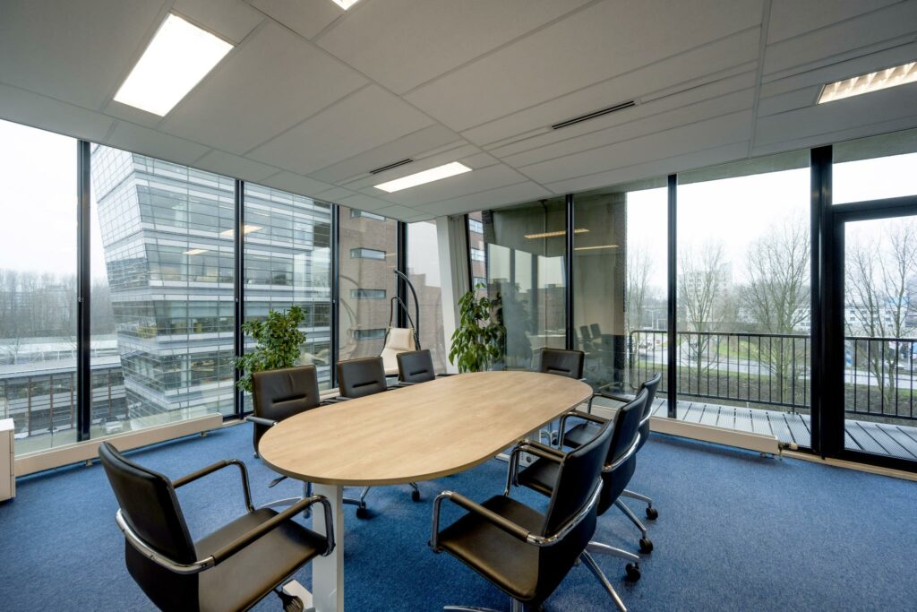 Vergaderruimte met ovale houten tafel, zwarte stoelen en uitzicht op kantoren aan de Joop Geesinkweg.